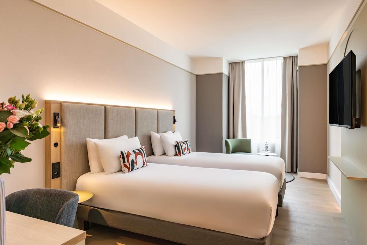 A hotel room with a bed, bedside table, chair, TV, and a window with curtains.