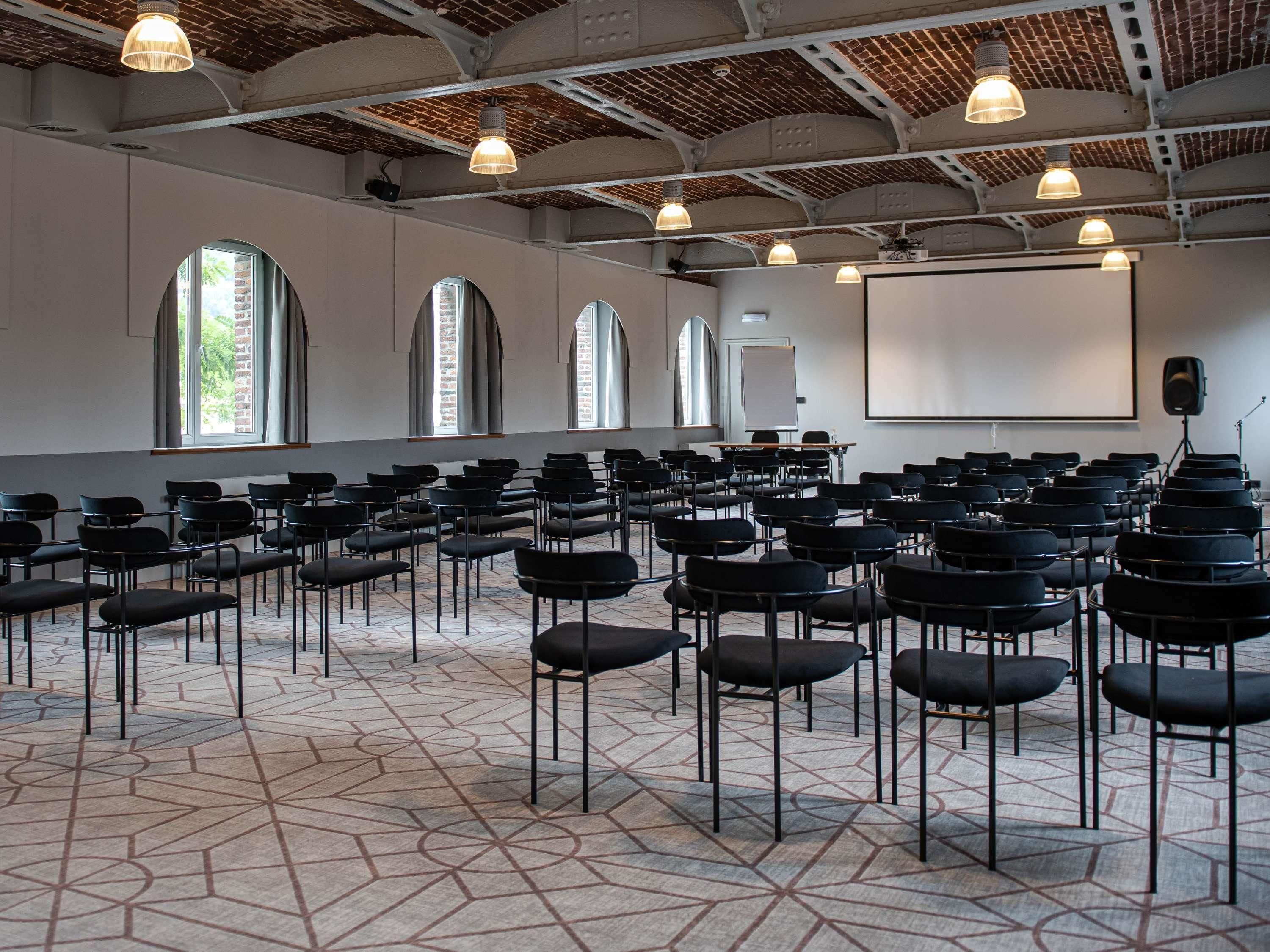 Une salle de conférence avec des rangées de chaises, un écran de projection et des fenêtres en arc de cercle.