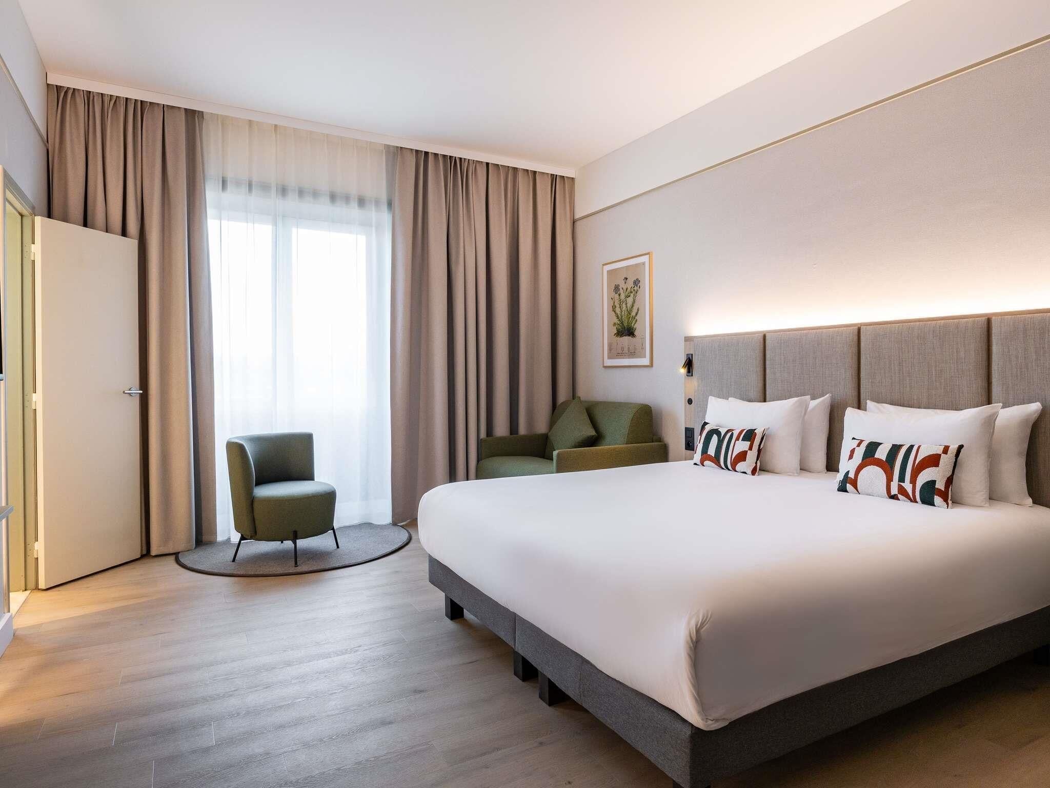 Quarto de hotel com uma cama grande, uma poltrona verde, um sofá e uma janela com cortinas.