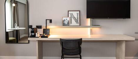 A modern hotel room with a large desk, a flat-screen TV mounted on the wall, a mirror, and a chair.