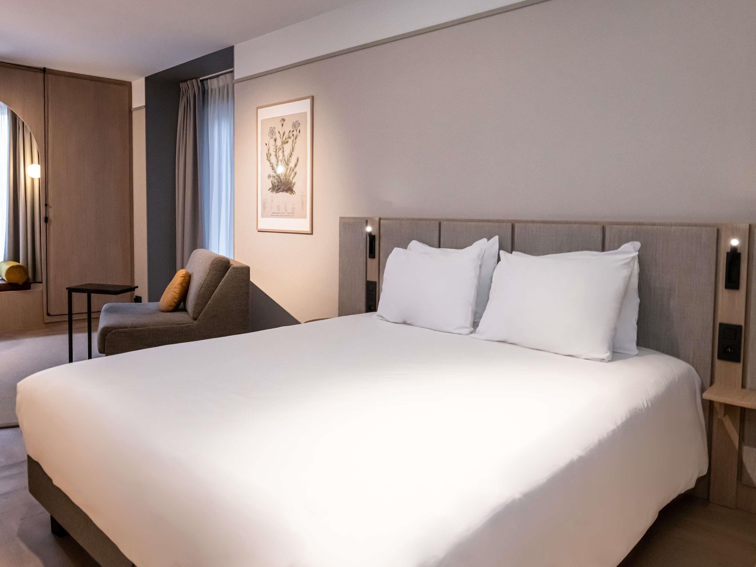 Een hotelkamer met een groot bed, een stoel, een klein tafeltje en een lijstje aan de muur.