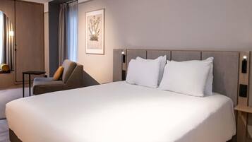 Een hotelkamer met een groot bed, een stoel, een klein tafeltje en een lijstje aan de muur.