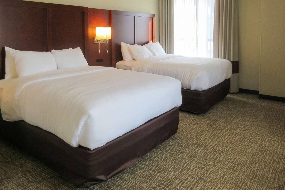 A hotel room with two beds, a wooden headboard, a wall-mounted lamp, and a window with curtains.
