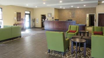 A hotel lobby with a reception desk, green armchairs, a small table, and a patterned rug.
