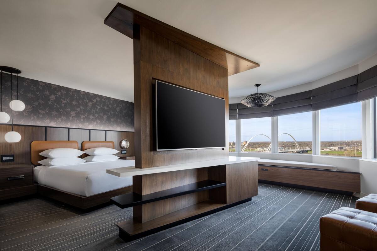 Une chambre d’hôtel moderne dotée d’une grande télévision à écran plat, d’un lit avec du linge de maison blanc, d’une tête de lit en bois et d’une grande fenêtre donnant sur la ville.