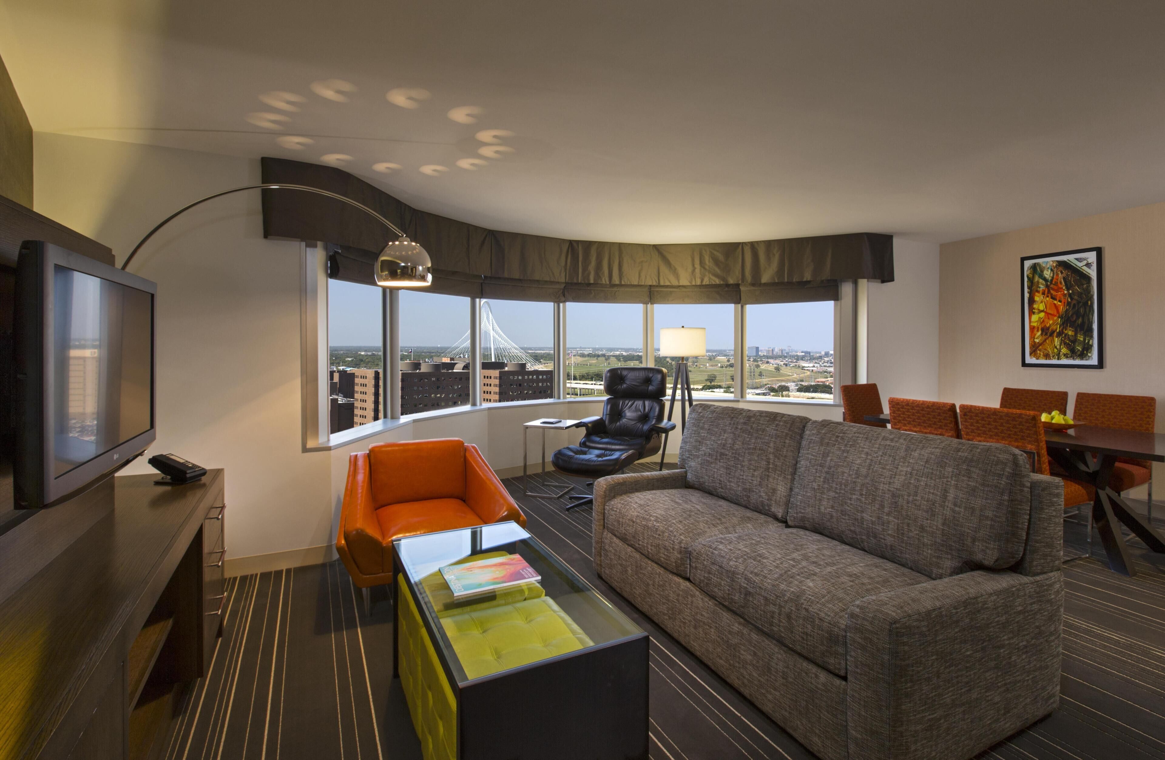 Una habitación de hotel moderna con un ventanal grande, un sofá, una mesa de centro, un televisor y un comedor con sillas.