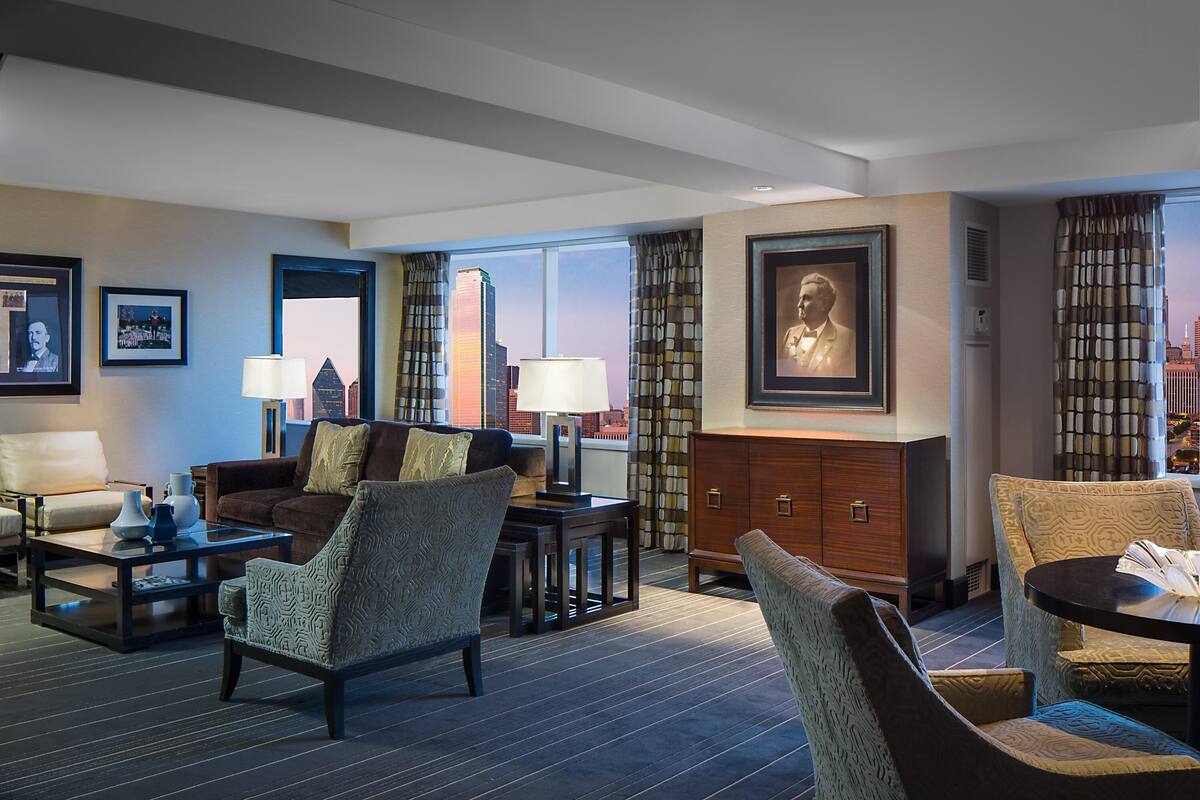 Une chambre d’hôtel avec un canapé, des fauteuils, une table basse, une lampe, un meuble et des tableaux encadrés aux murs.