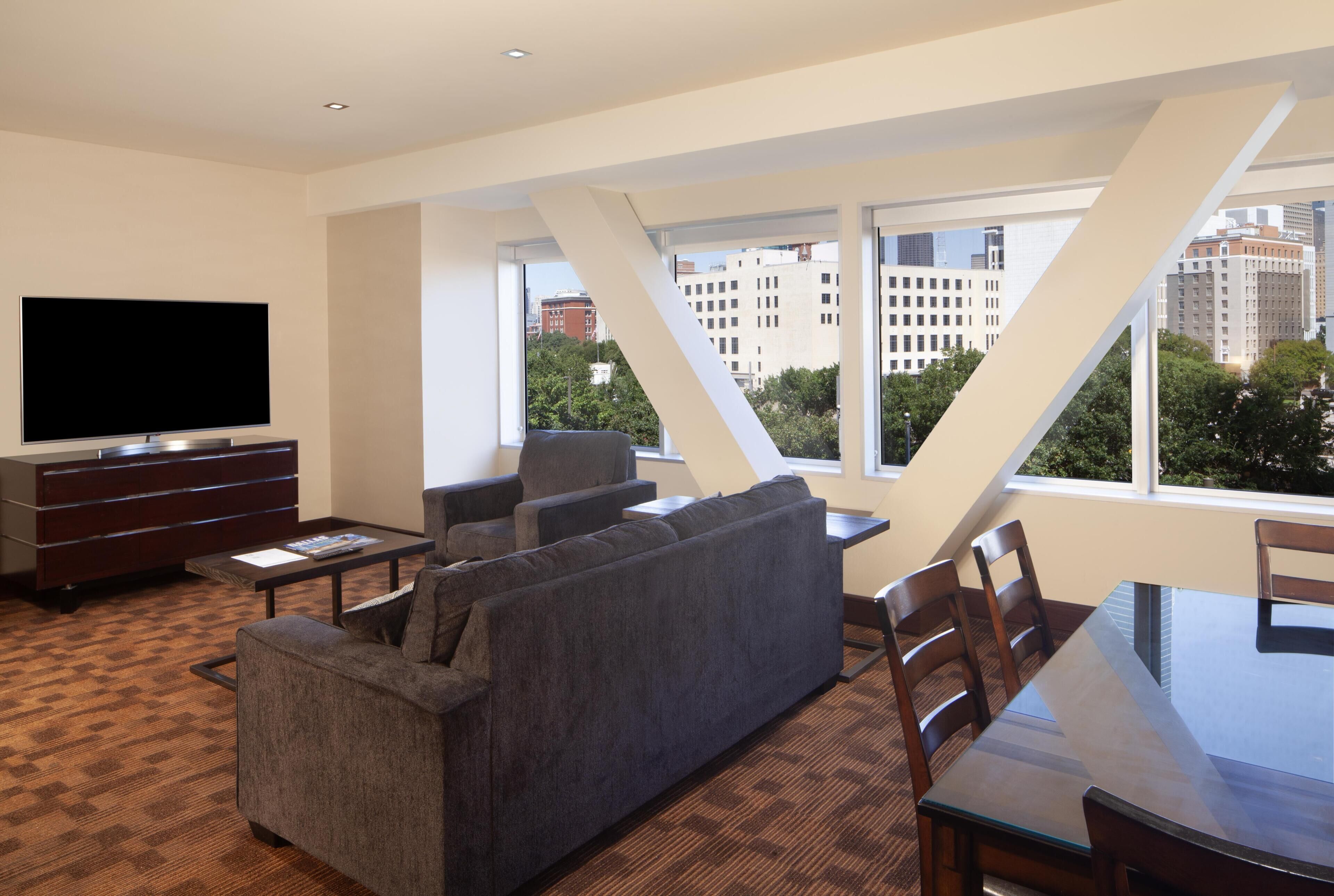 A modern living room with a flat-screen TV, a sofa, a dining table, and large windows offering a city view.