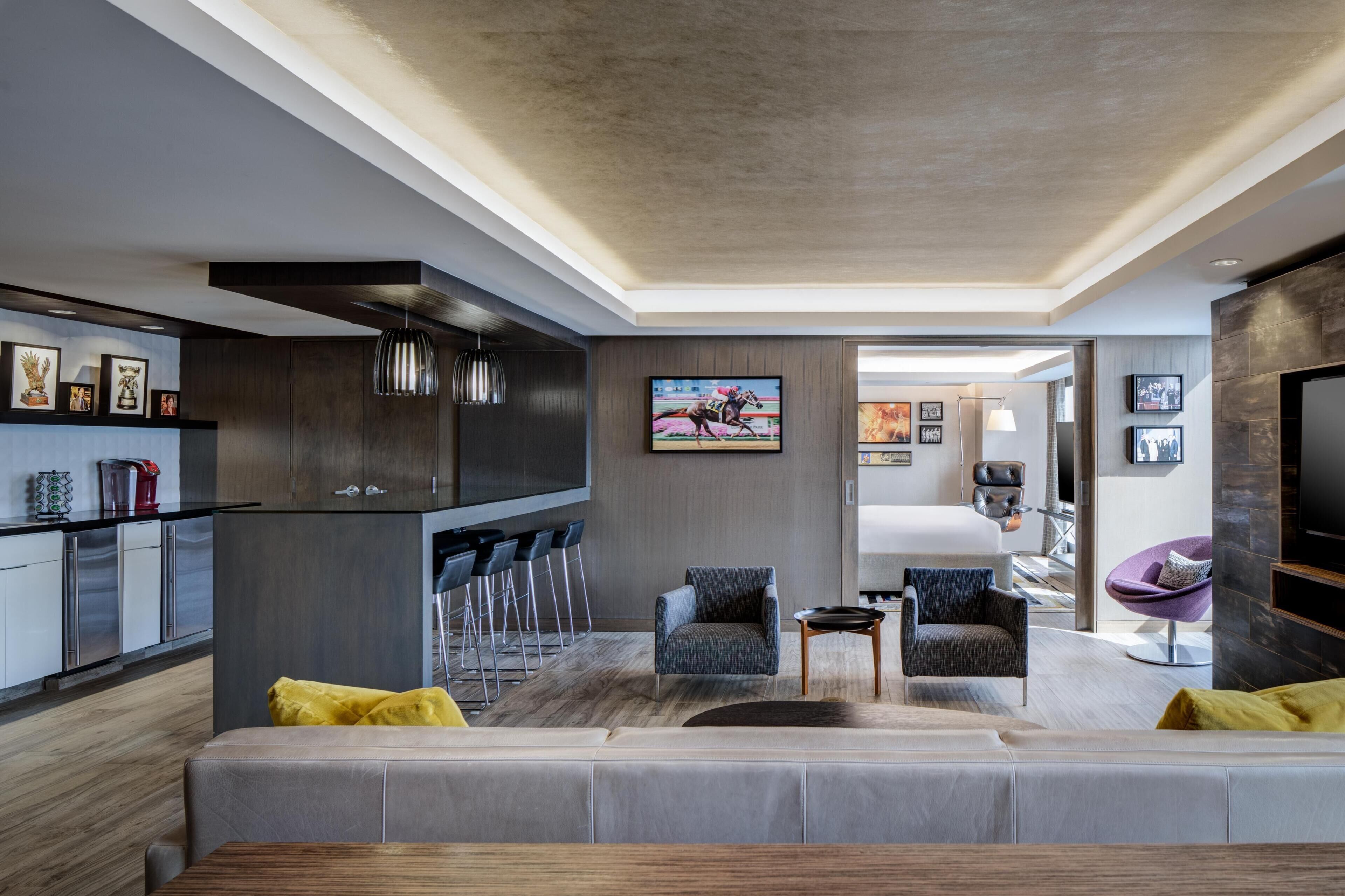 A modern living room with a grey sofa, a small coffee table, and a TV mounted on the wall. There is a kitchen area with a bar and stools, and a dining table with chairs.