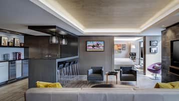Uma sala de estar moderna com um sofá cinzento, uma pequena mesa de centro e uma televisão fixada na parede. Há uma área de cozinha com um balcão e banquetas, e uma mesa de jantar com cadeiras.
