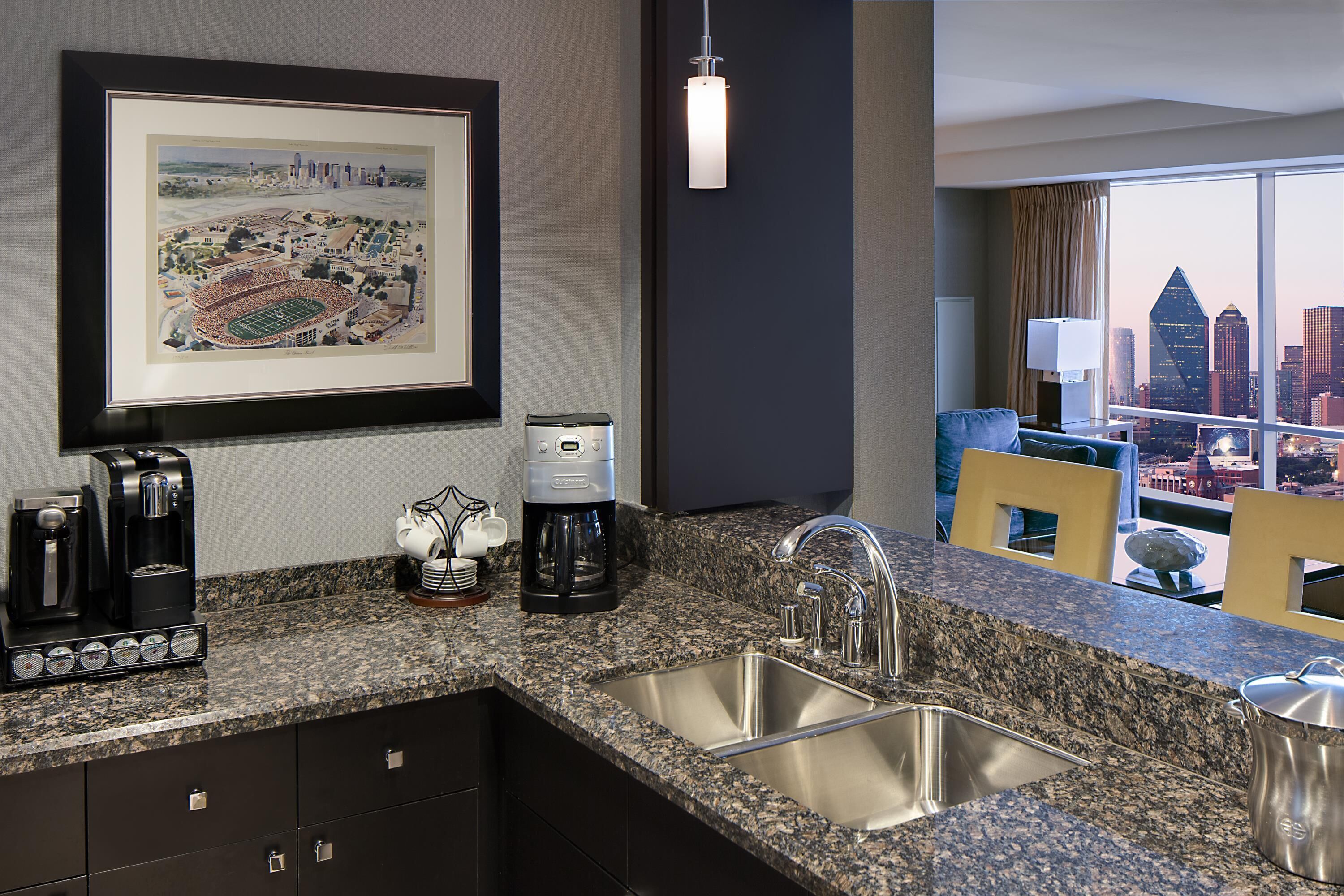 A hotel room with a kitchen area, a framed cityscape artwork, a coffee maker, and a view of the city skyline.