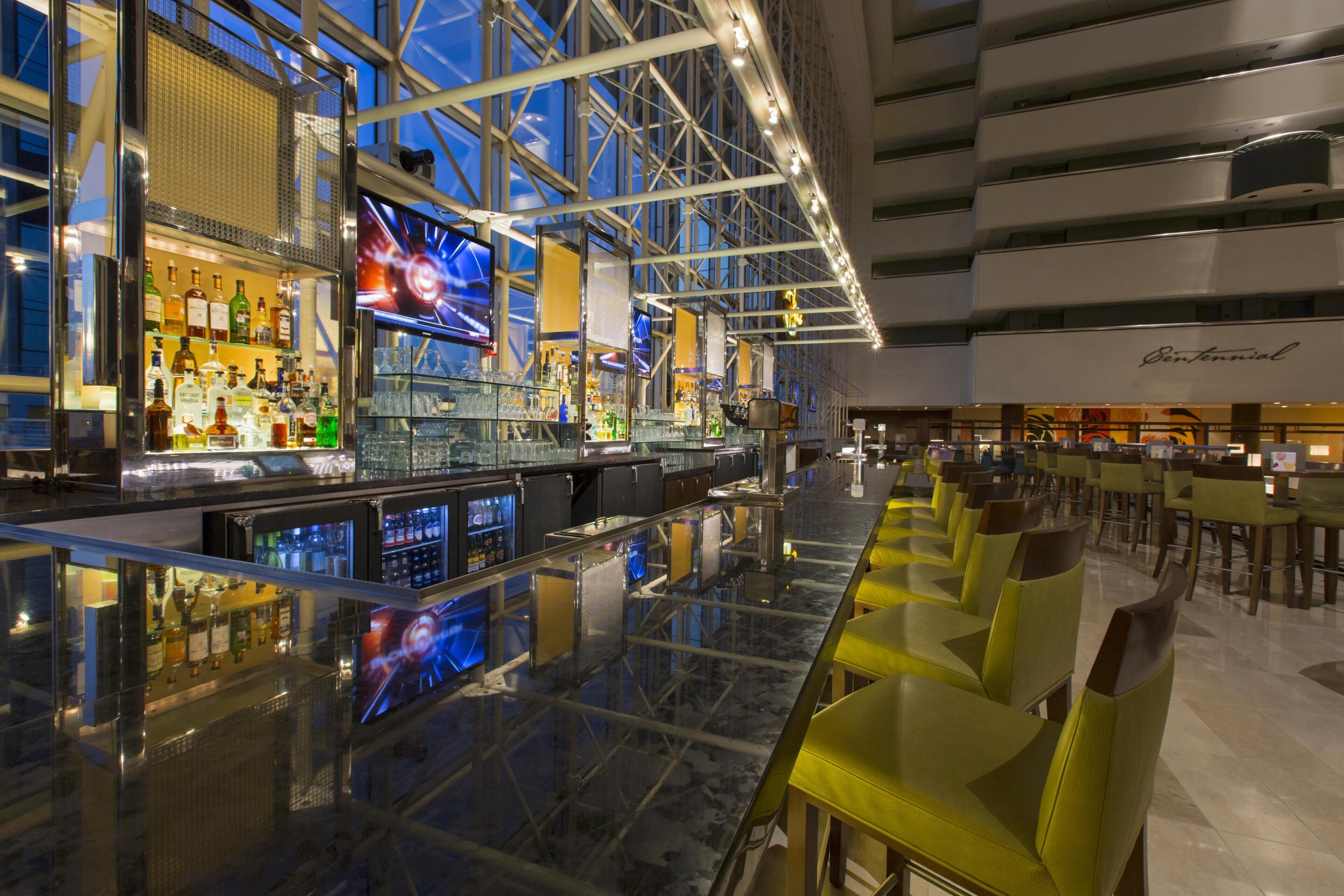 Uma área de bar moderna com balcão de vidro, várias telas e um espaço de estar com cadeiras amarelas.