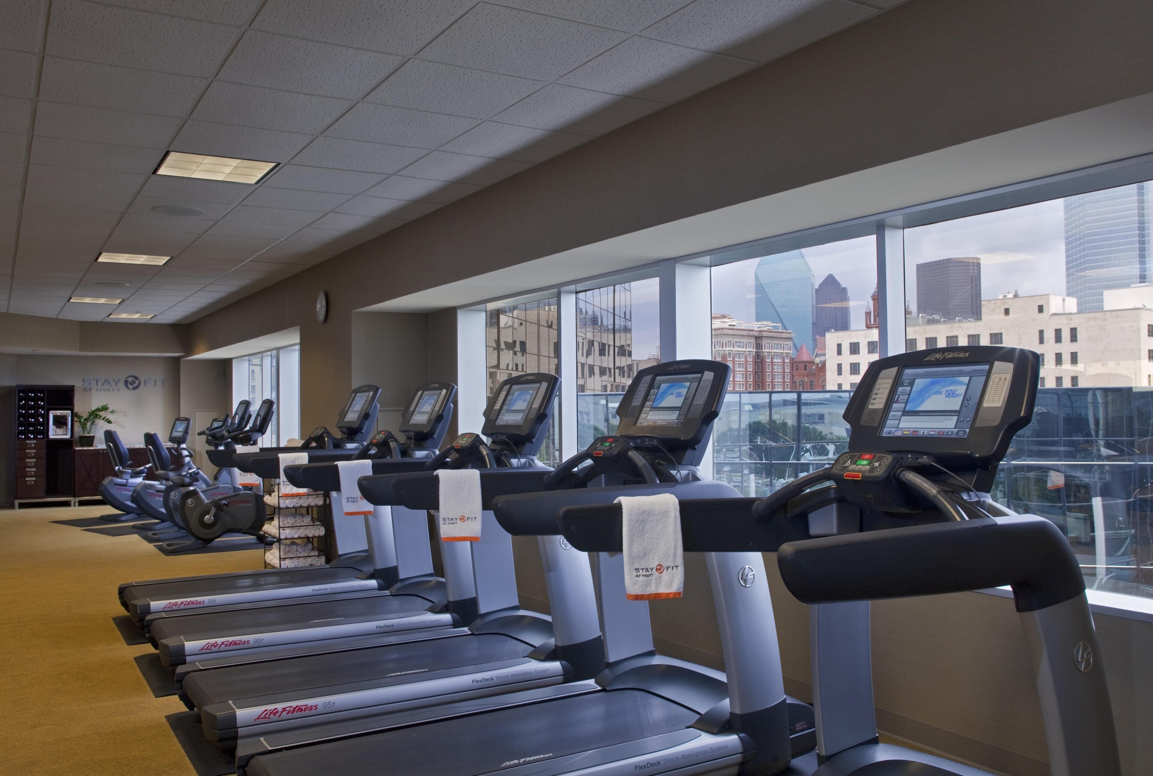 A row of treadmills with digital displays, a clock, and a 'Stay Fit' sign in a gym with large windows overlooking a cityscape.