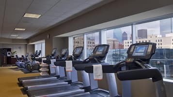 A row of treadmills with digital displays, a clock, and a 'Stay Fit' sign in a gym with large windows overlooking a cityscape.
