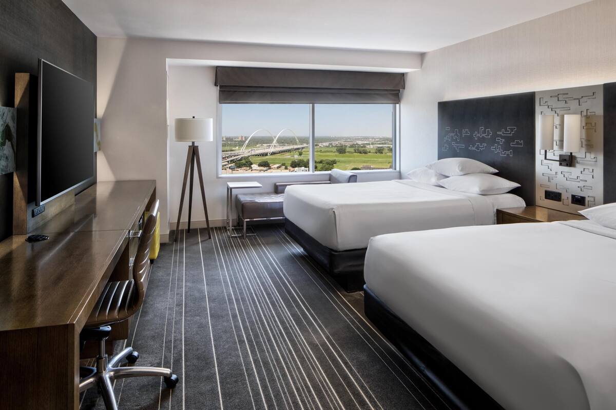 Une chambre d’hôtel avec une grande fenêtre, deux lits, un bureau avec une chaise, une lampe et un téléviseur.