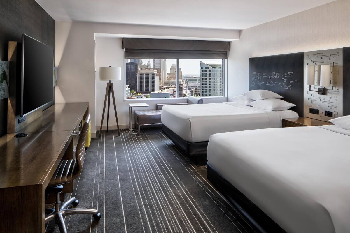 Une chambre d’hôtel avec vue sur la ville, un bureau avec un téléviseur à écran plat, deux lits, une lampe et un mur orné d’un motif géométrique.