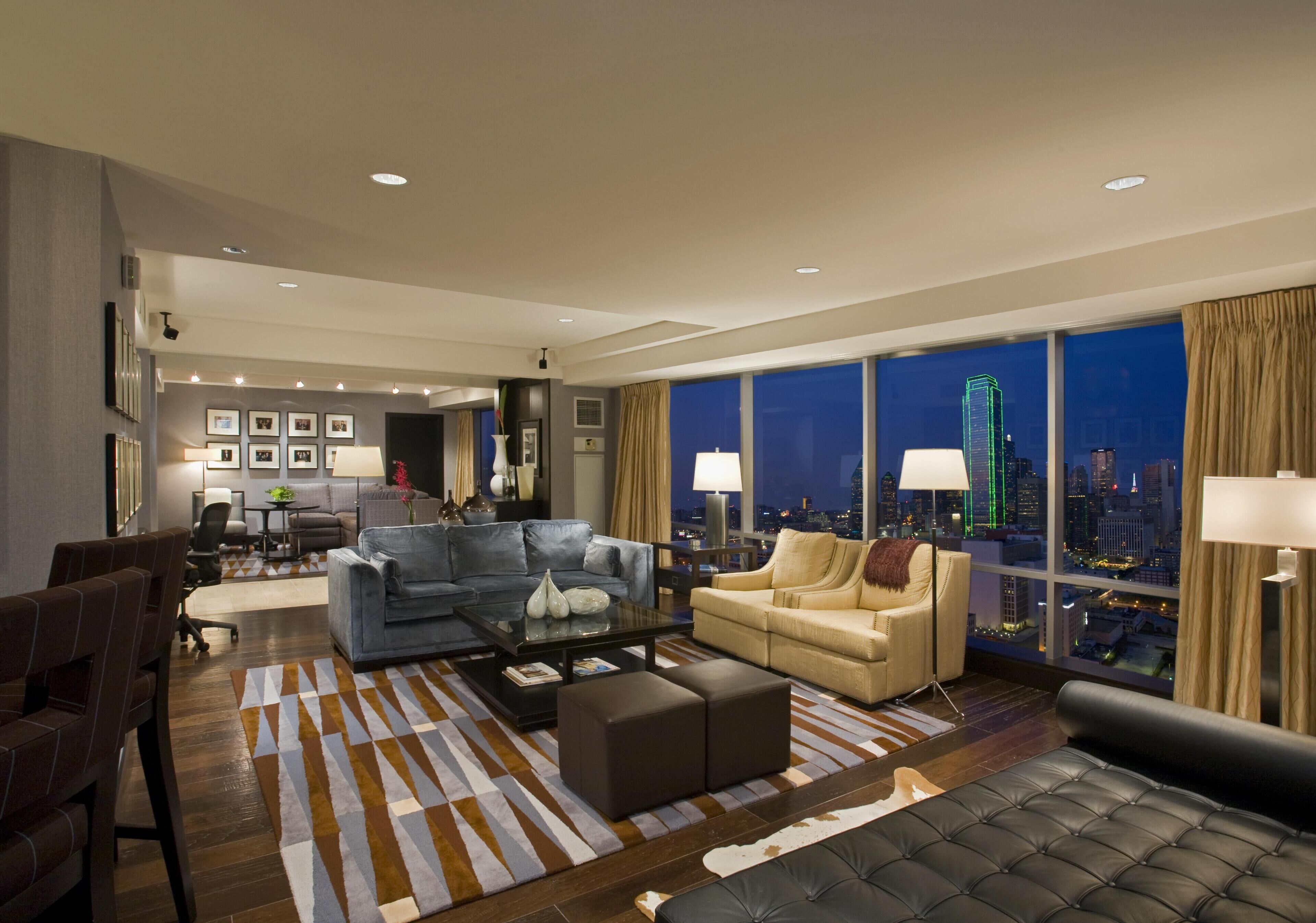 A modern hotel room with a city view, a sofa, a coffee table, and a dining area.