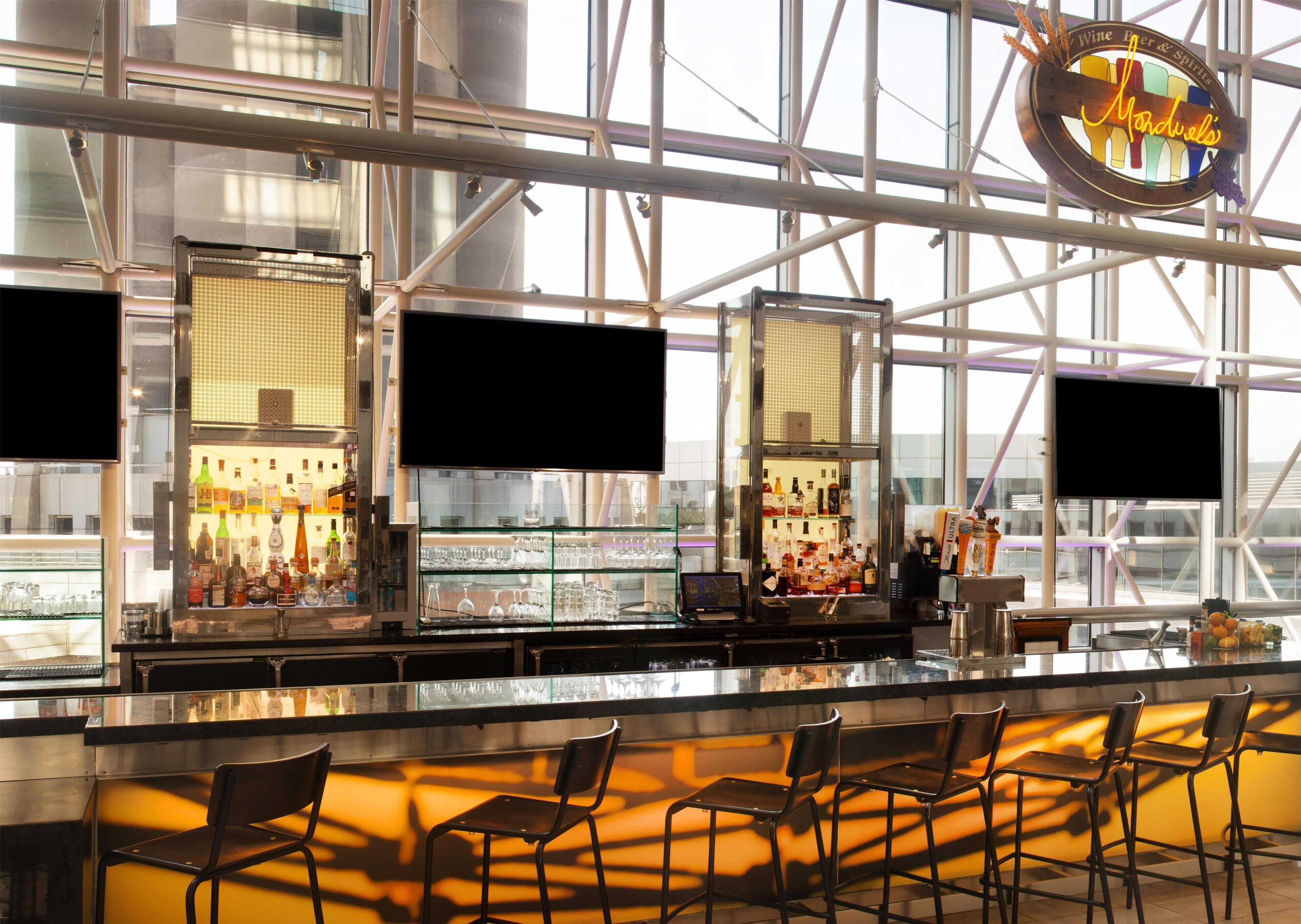 A bar area with a modern design, featuring a glass partition, bar stools, and a variety of bottles and glasses.