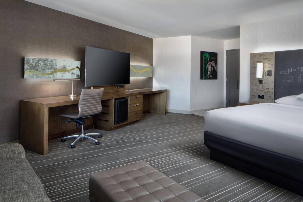 Une chambre d’hôtel comprenant un lit, un bureau avec une chaise, une télévision, des œuvres d’art accrochées aux murs et une porte de salle de bain.