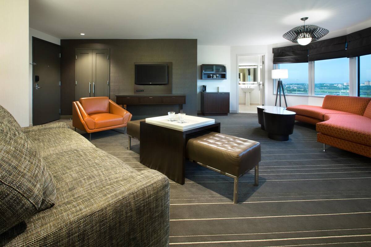 Une chambre d’hôtel moderne dotée d’un canapé, d’un fauteuil, d’un pouf, d’une télévision à écran plat et offrant une vue sur la ville.