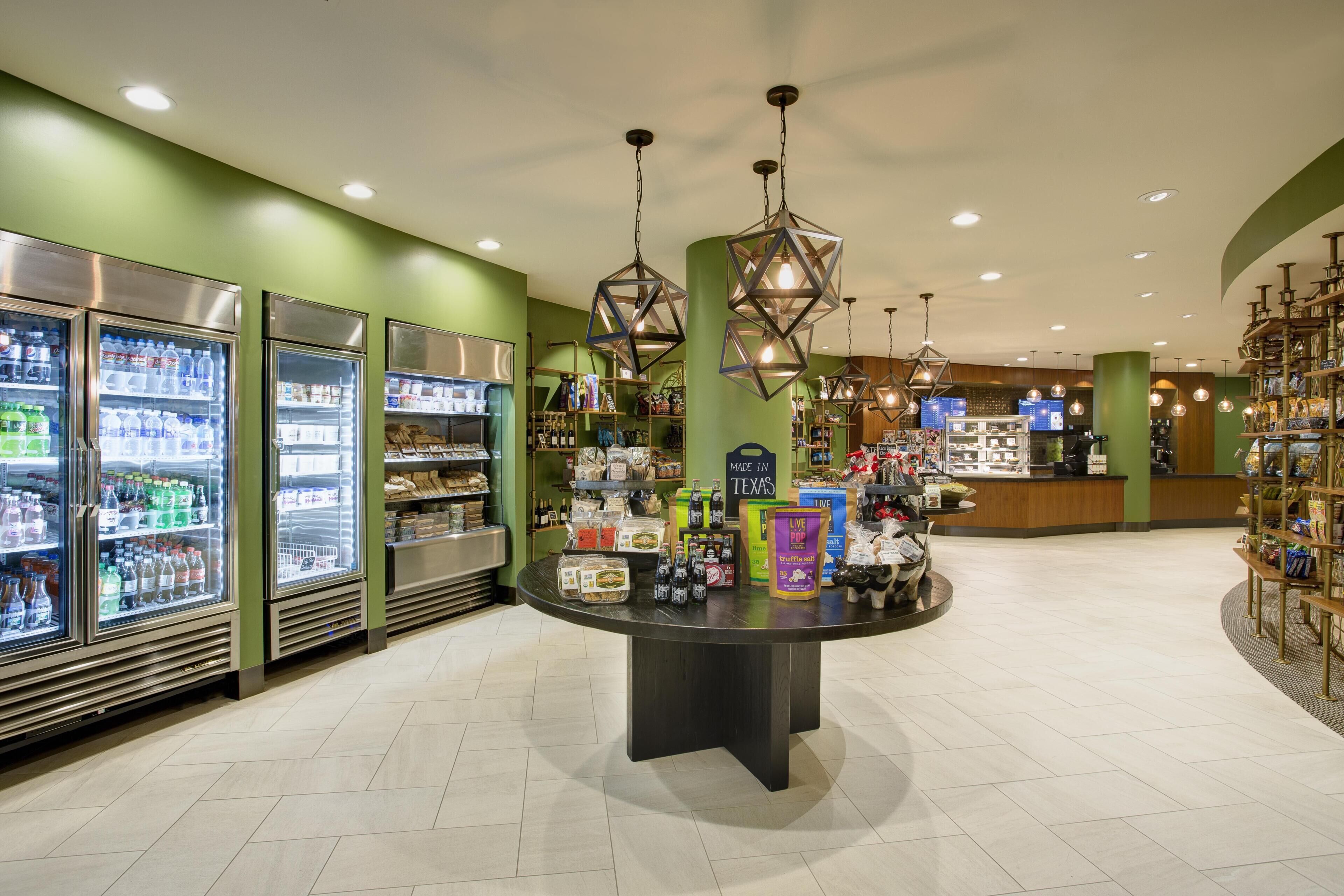 A well-lit store with refrigerated beverage dispensers, a central display table with various products, and shelves stocked with snacks and beverages.