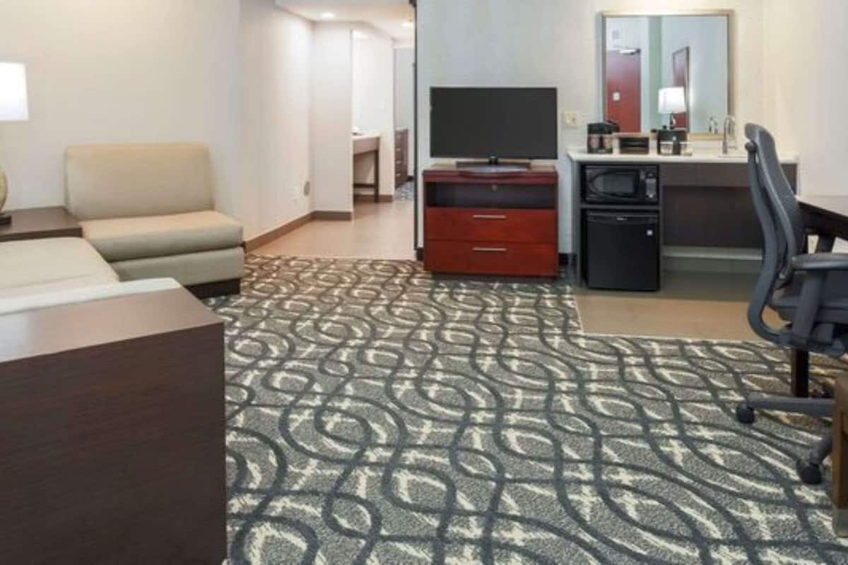 Habitación de hotel con sofá, televisión, escritorio con computadora, kitchenette pequeña y alfombra estampada.