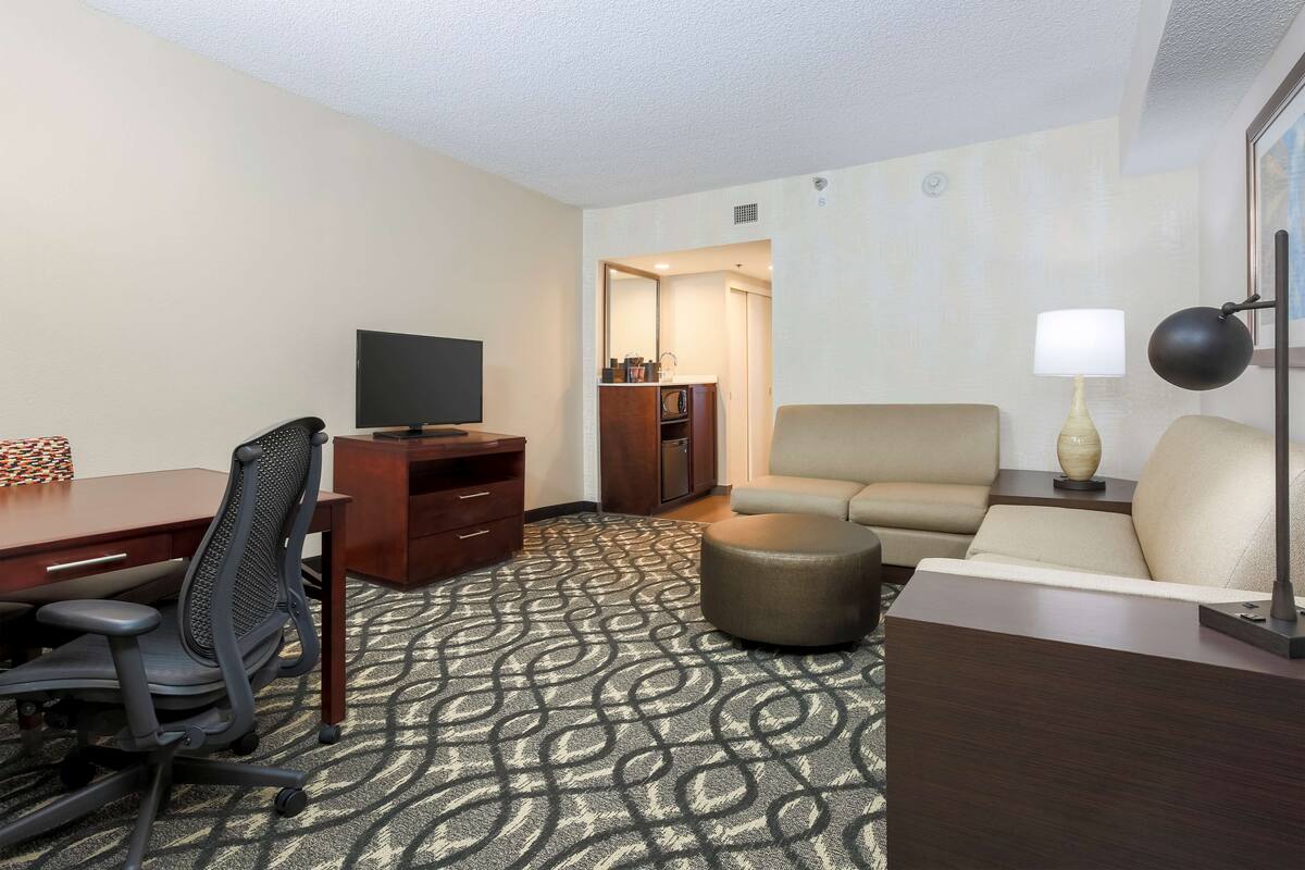 Una habitación de hotel con escritorio, silla, televisor, sofá, otoman y lámpara.