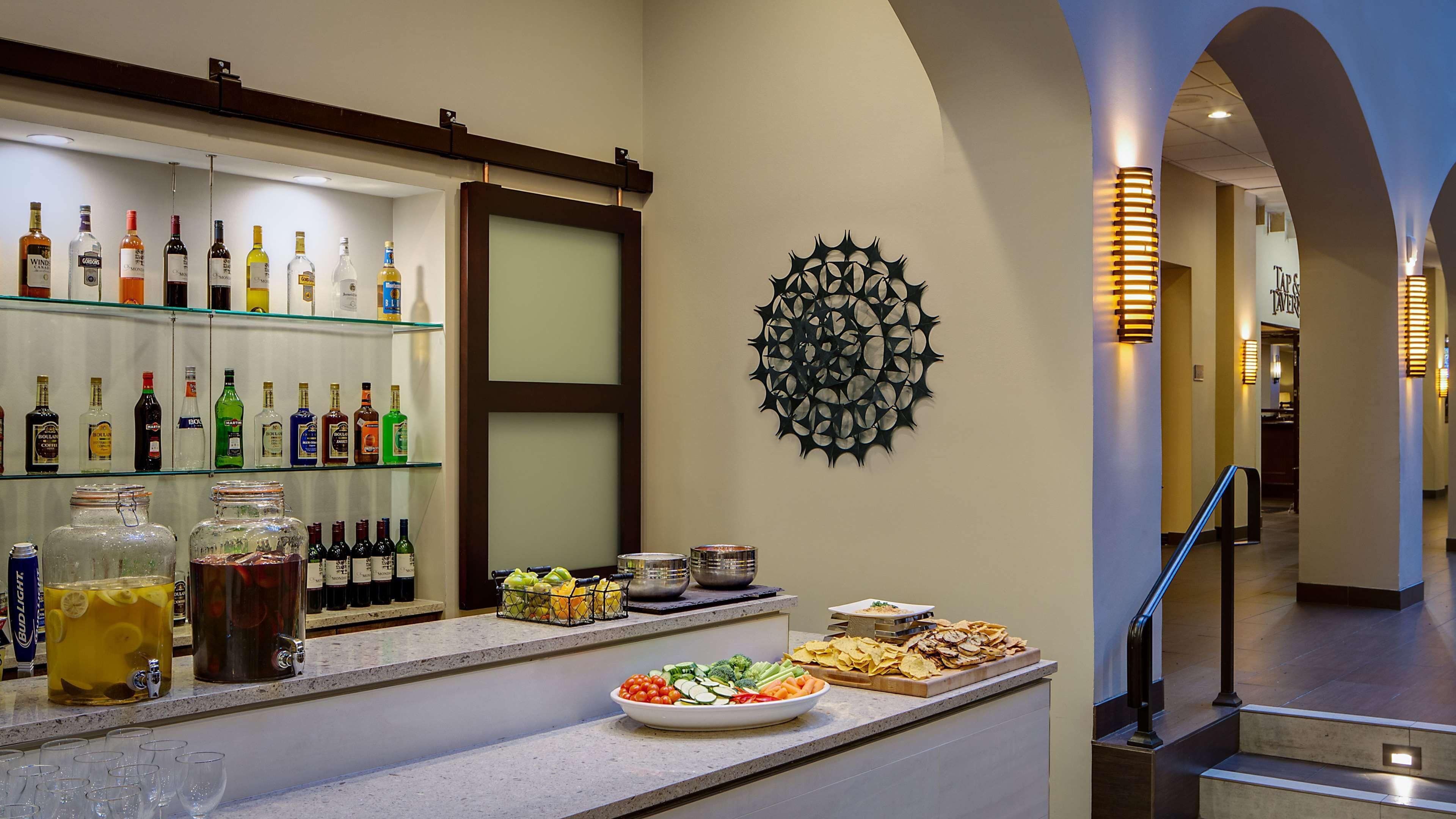 Un espace bar avec différentes bouteilles d’alcool, un distributeur de punch aux fruits et un élément décoratif mural.