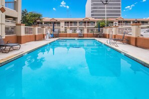 Una piscina rectangular con superficie de agua azul cristalina, rodeada por un muro de ladrillo bajo y una zona cubierta con sillas y mesas.