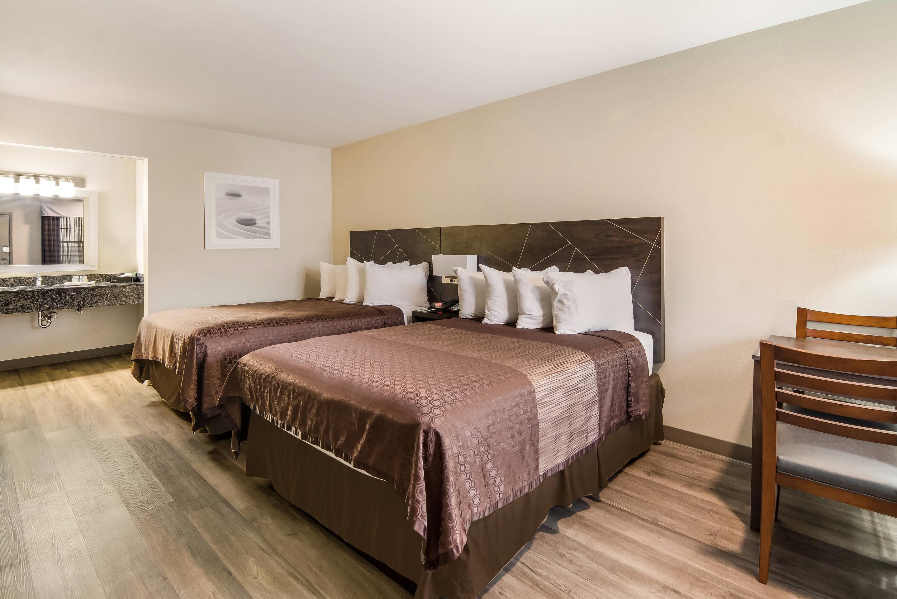 Quarto de hotel com duas camas, uma cadeira de madeira, banheiro com espelho e um quadro decorativo na parede.