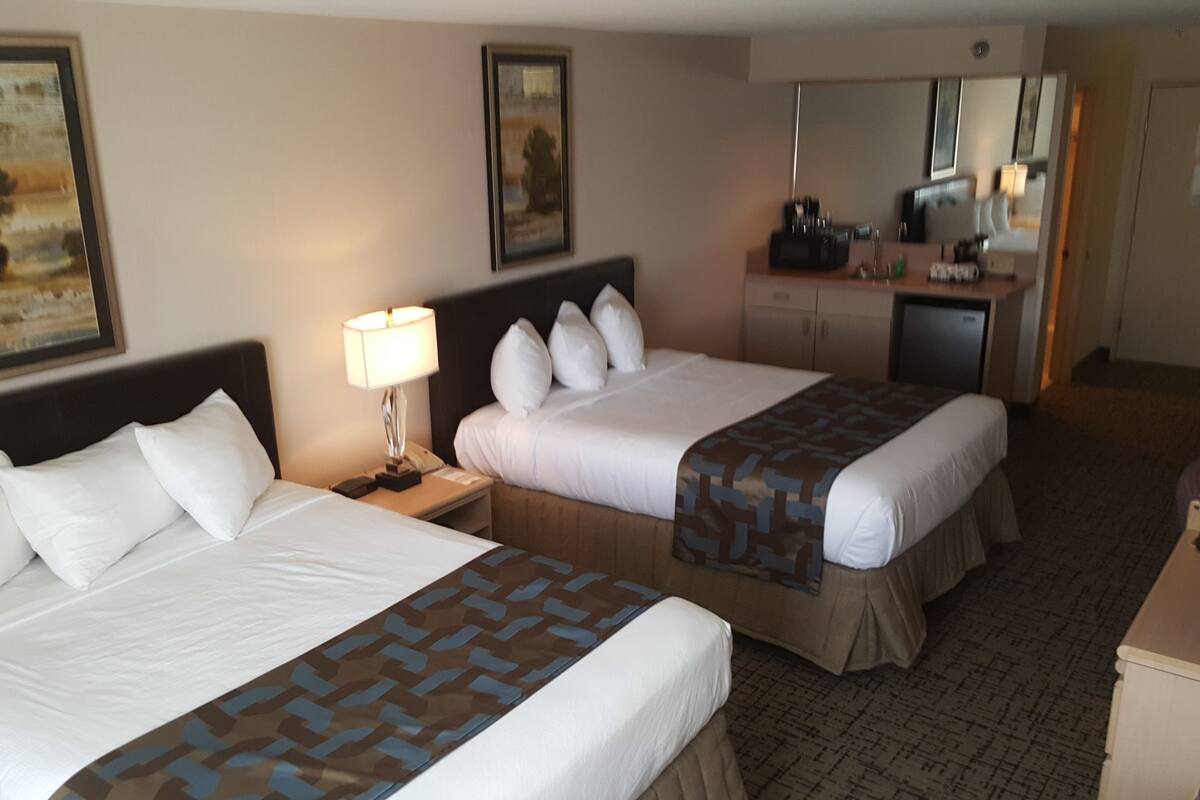 A hotel room with two beds, a desk, a lamp, a refrigerator, and a mirror.