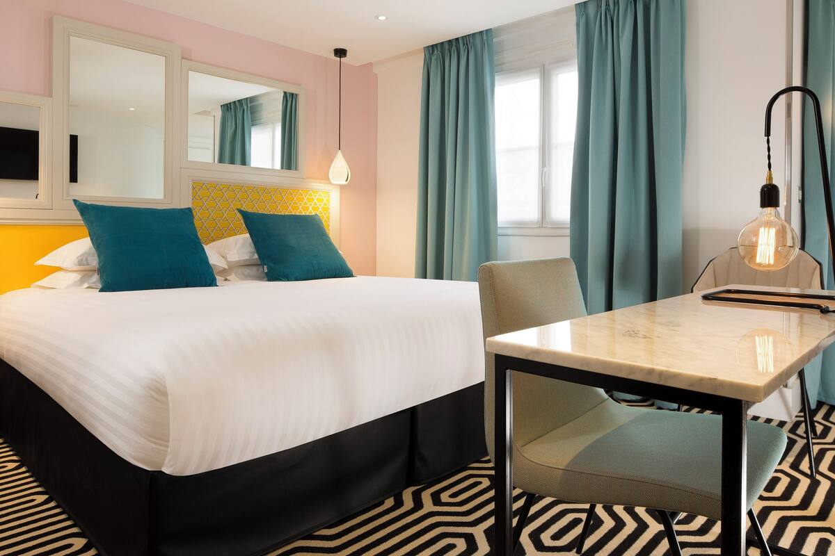 A hotel room with a bed, a table, a chair, a desk lamp, and a patterned floor.