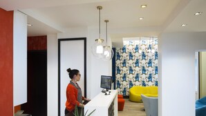 A reception area with a receptionist, a computer monitor, a plant, and colorful seating. - Hôtel Haussmann Saint Augustin (Paris)
