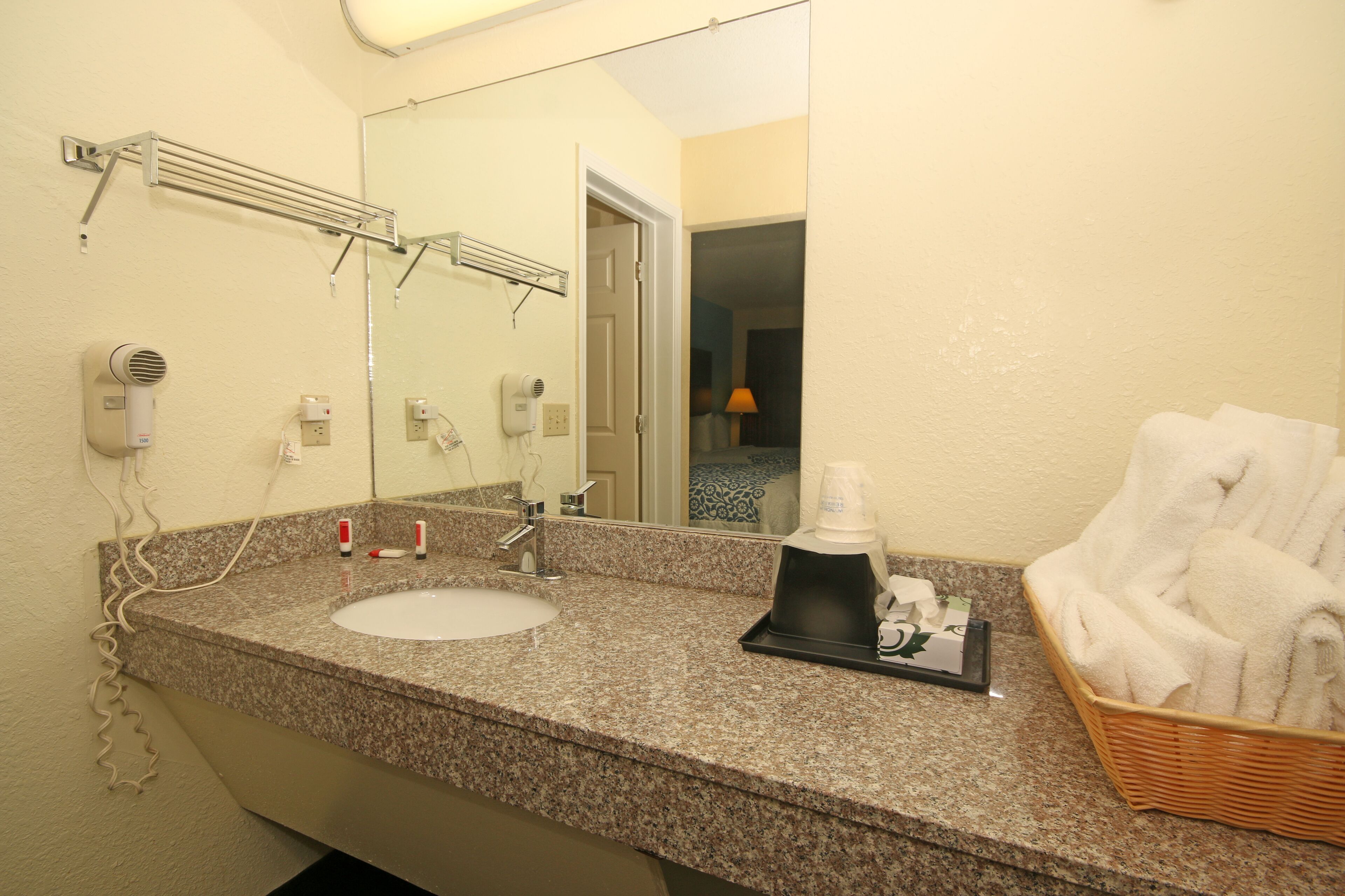 Une salle de bain dotée d’un comptoir en granit, d’un lavabo, d’un miroir, d’un sèche-cheveux, d’un porte-serviettes et d’un panier rempli de serviettes.
