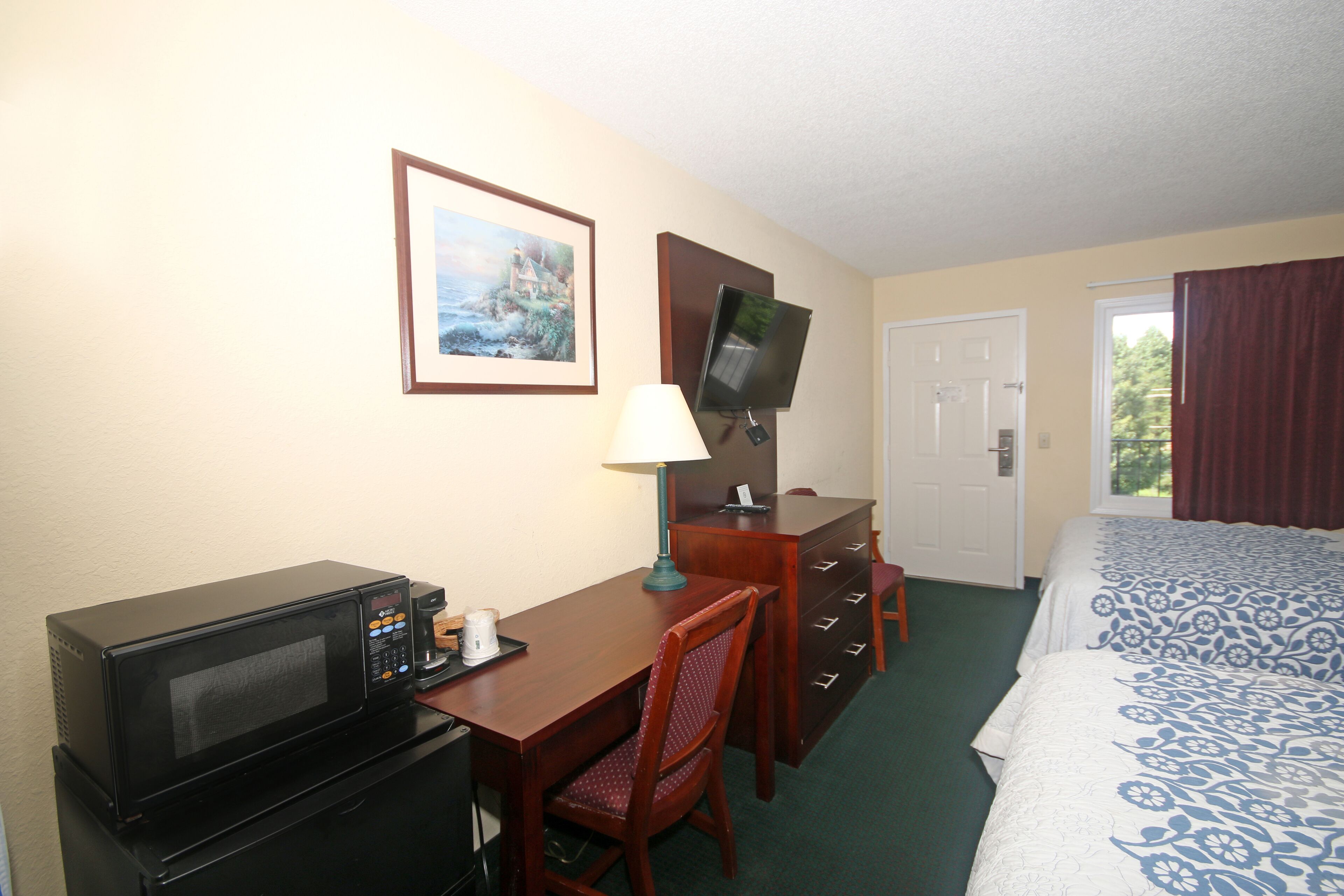 A hotel room with a bed, a desk with a microwave, a chair, a dresser, a television, and a framed picture on the wall.