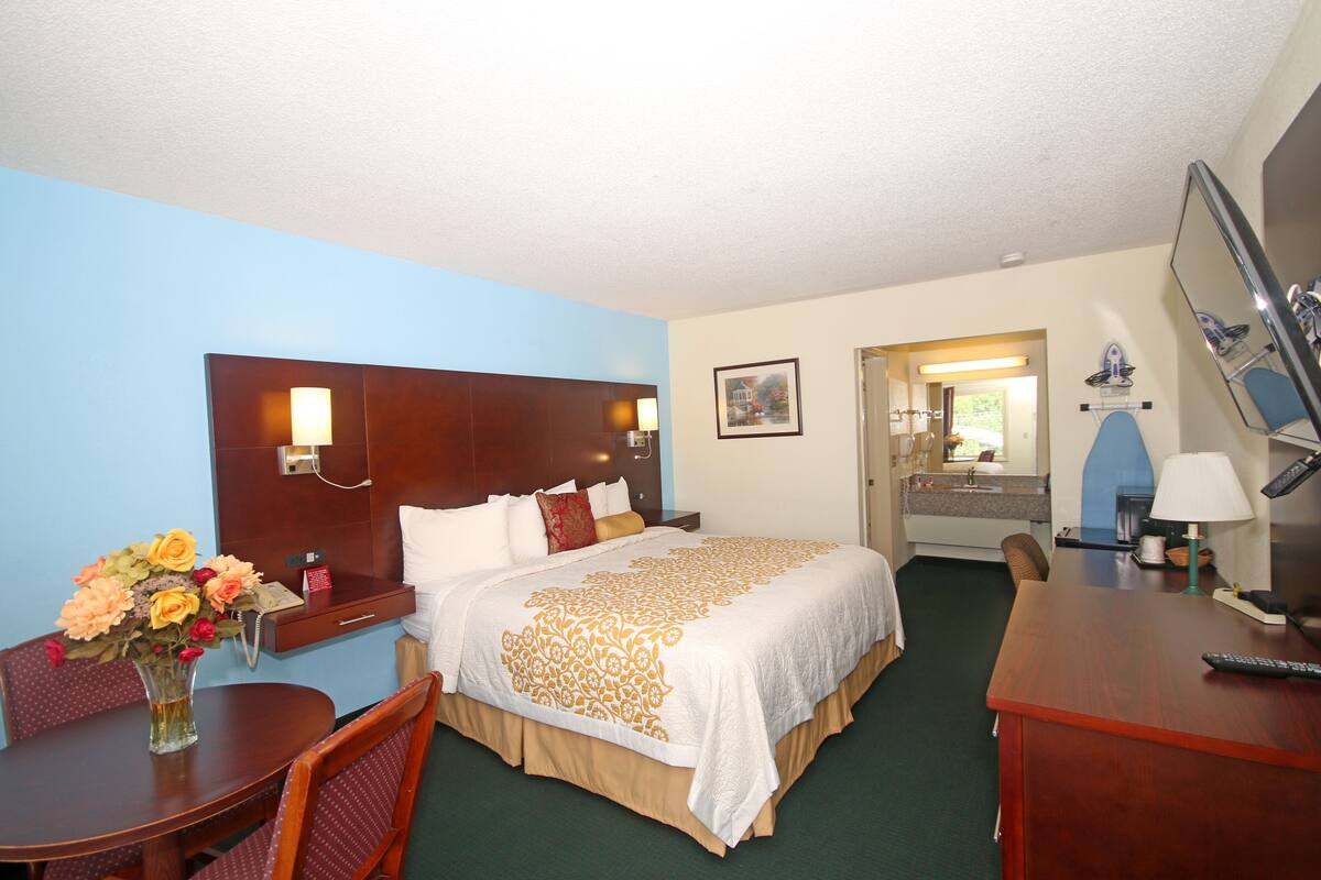 A hotel room with a large bed, a desk, a chair, a small table with a vase of flowers, and a bathroom visible through an open door.