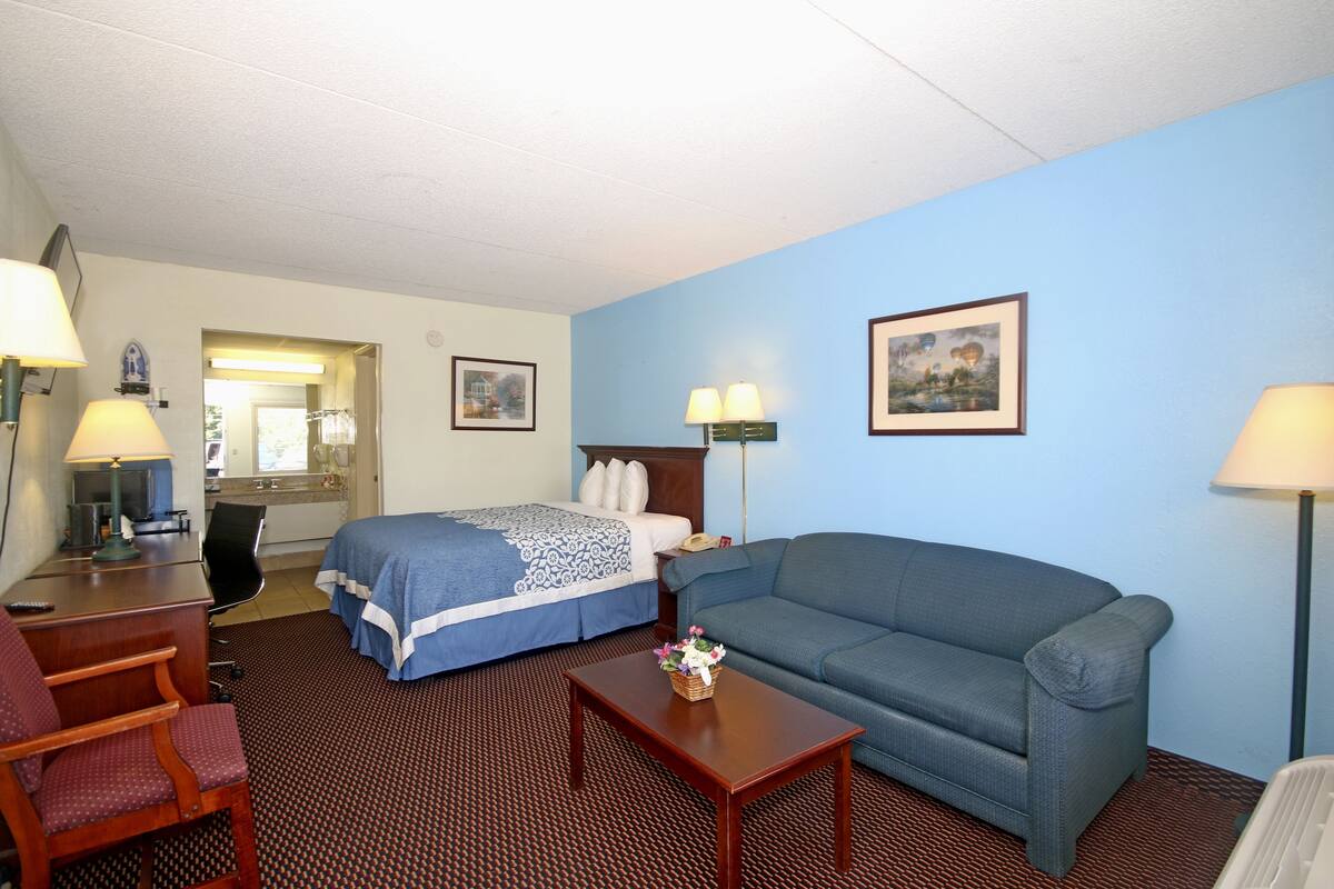 A hotel room with a bed, a blue sofa, a wooden coffee table, a desk with a lamp, a chair, and two framed pictures on the wall.