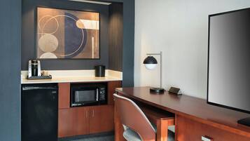 A compact hotel room with a kitchenette, a desk, a chair, a flat-screen TV, and a wall-mounted abstract painting.