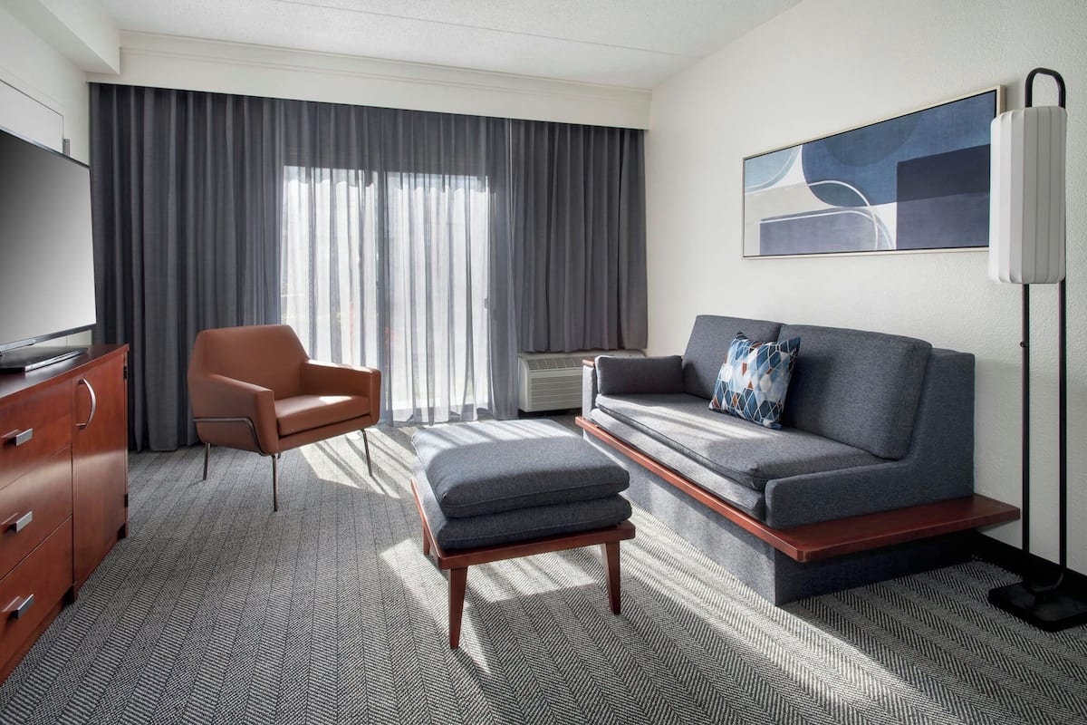 A modern hotel room with a sofa, armchair, ottoman, television, and artwork on the wall.