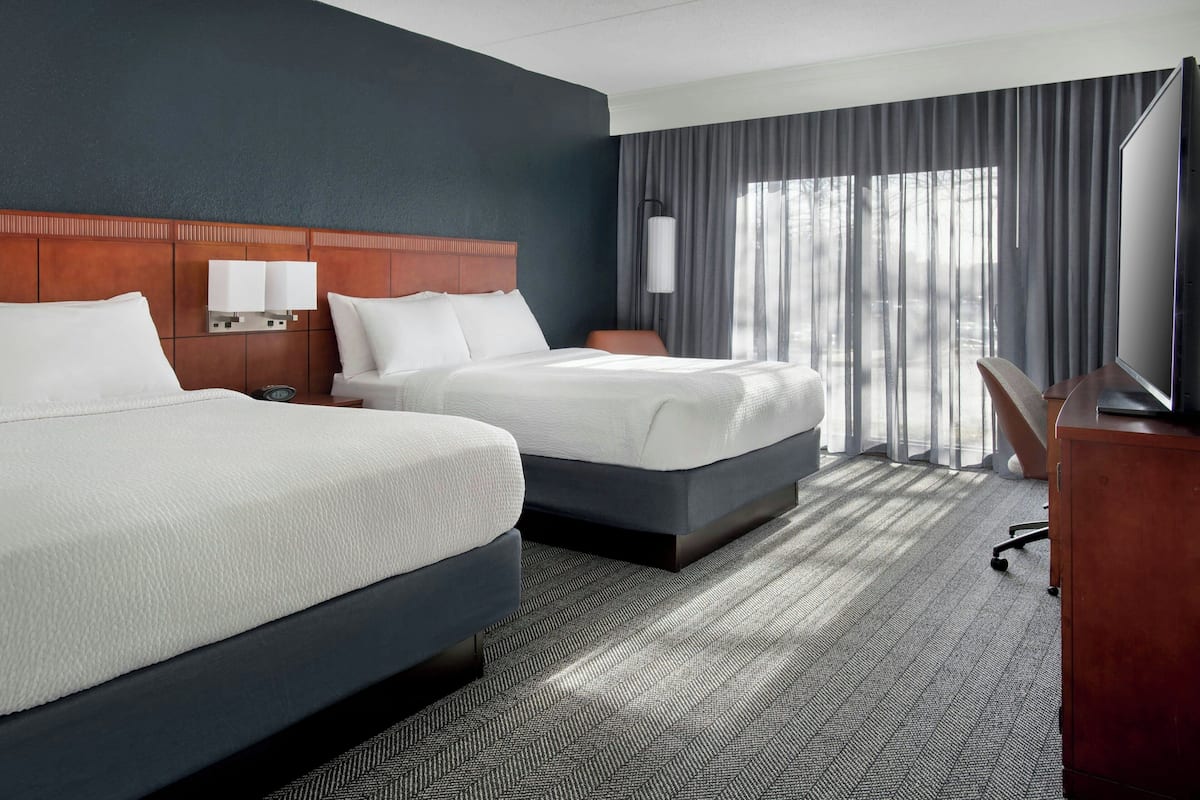 A hotel room with two beds, a wooden headboard, a desk with a chair, and a large window with curtains.