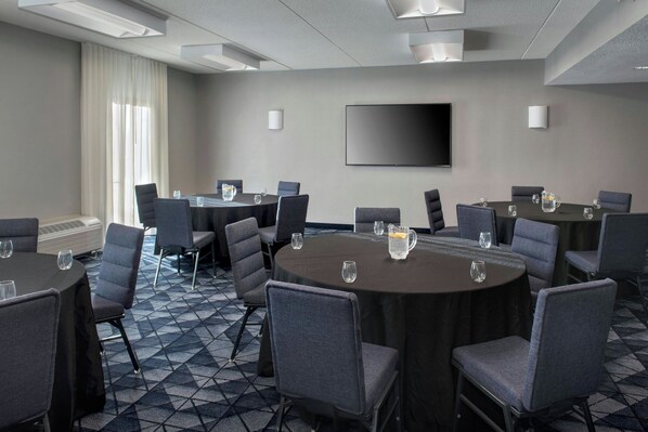 A conference room with round tables, gray chairs, a flat-screen TV, and a large window with curtains.