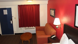 A hotel room with a red wall, a wooden chair, a sofa, a bed, a lamp, and a window with red curtains. - Americas Best Value Inn Fredericksburg N (Fredericksburg)