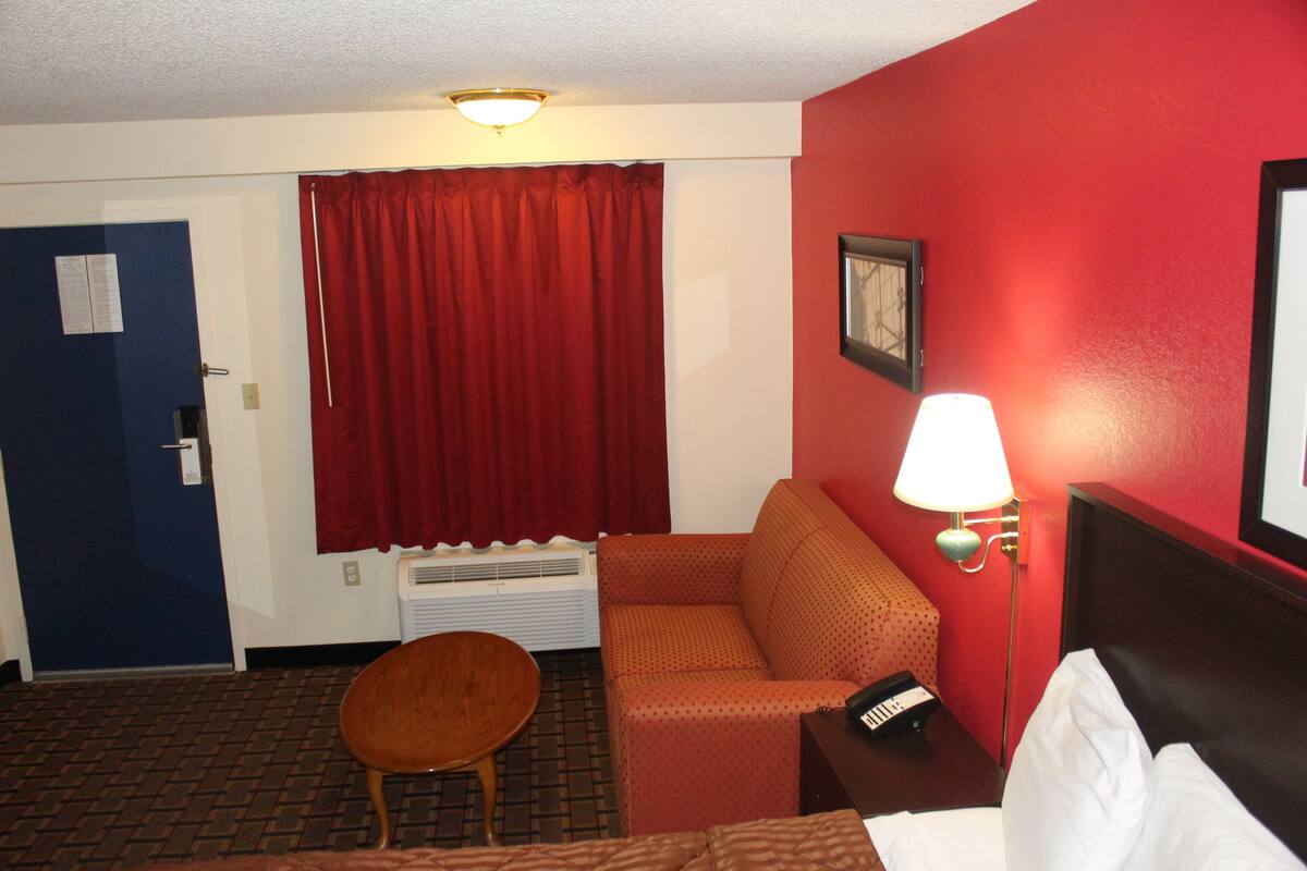 A hotel room with a red wall, a wooden chair, a sofa, a bed, a lamp, and a window with red curtains.