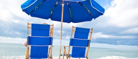 Two beach chairs under a blue umbrella on a snowy beach.