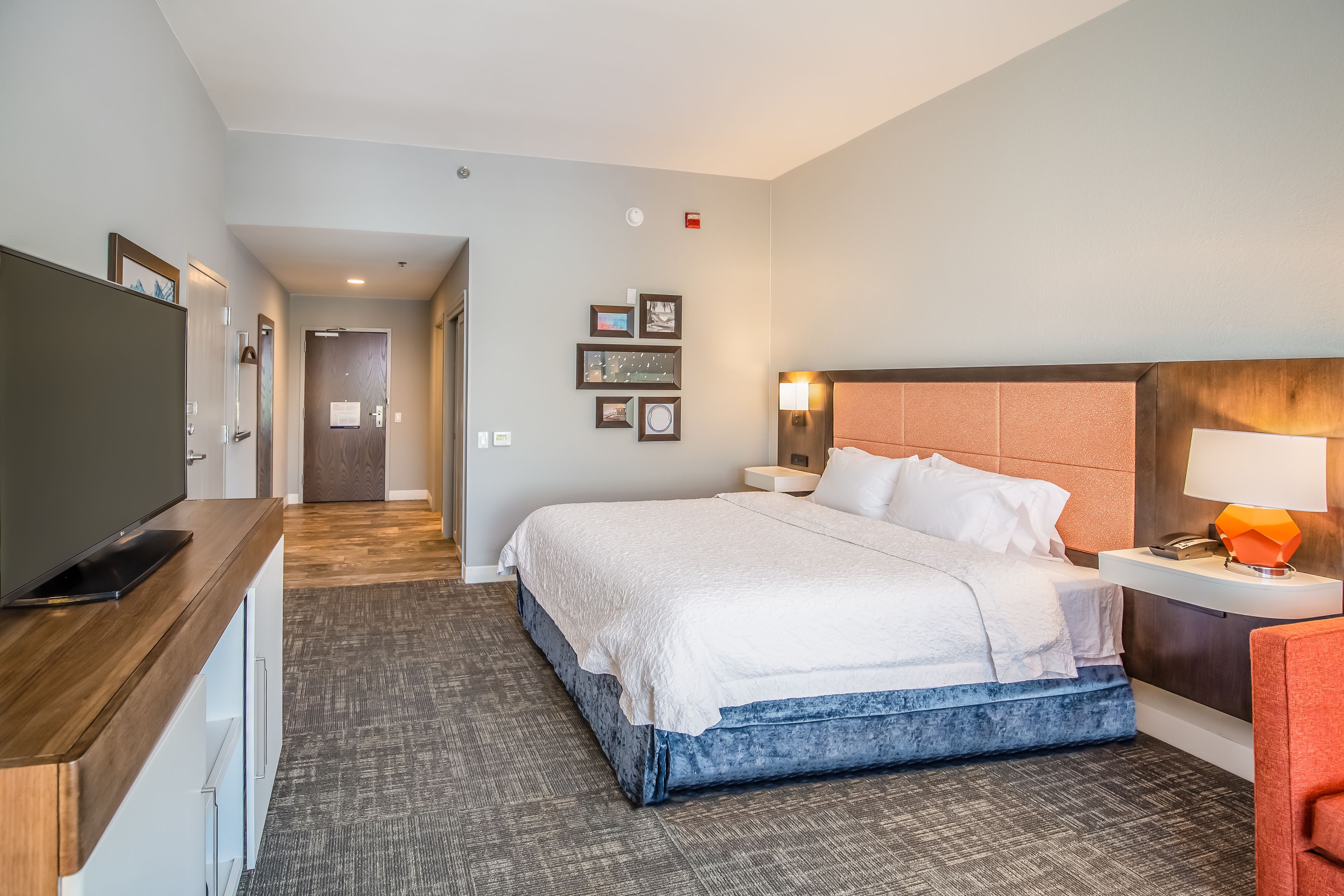 A hotel room with a large bed, a desk with a flat-screen TV, a bedside table with a lamp, and a wall-mounted TV.