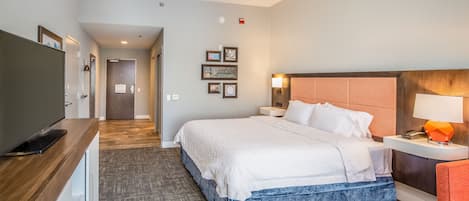 A hotel room with a large bed, a desk with a flat-screen TV, a bedside table with a lamp, and a wall-mounted TV.