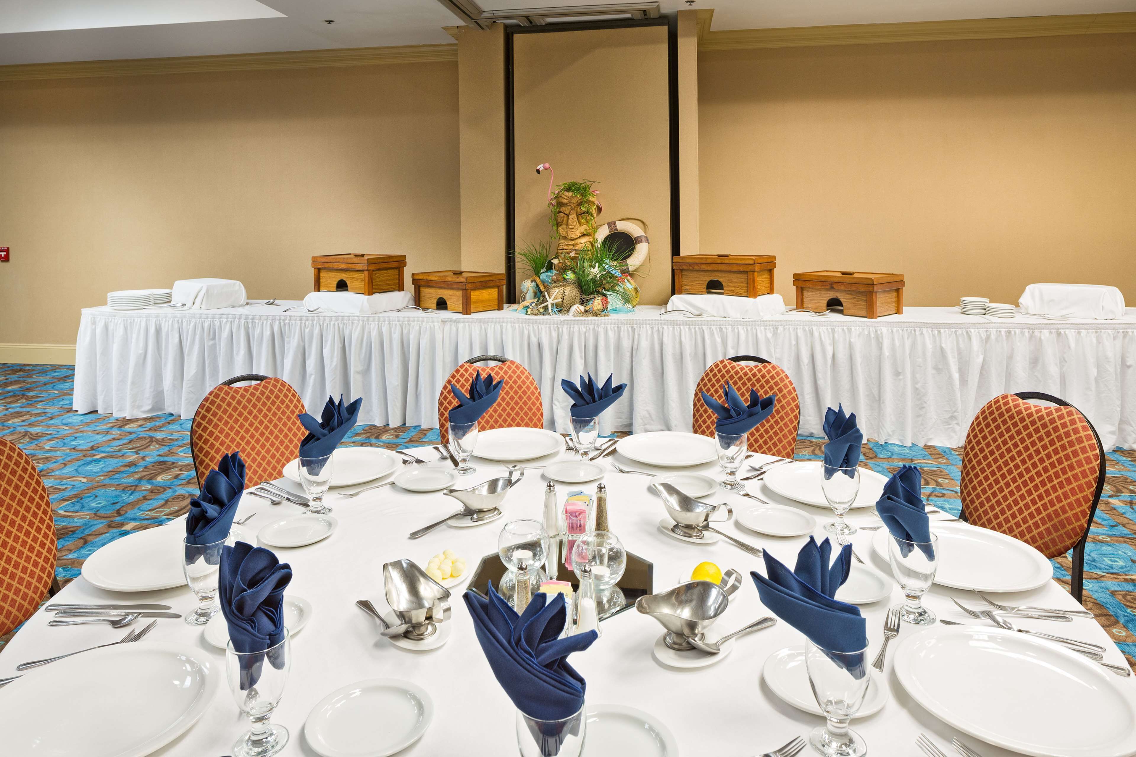 Un salón de conferencias con una larga mesa dispuesta para una reunión, equipado con sillas, platos, cubiertos y un centro de mesa decorativo.