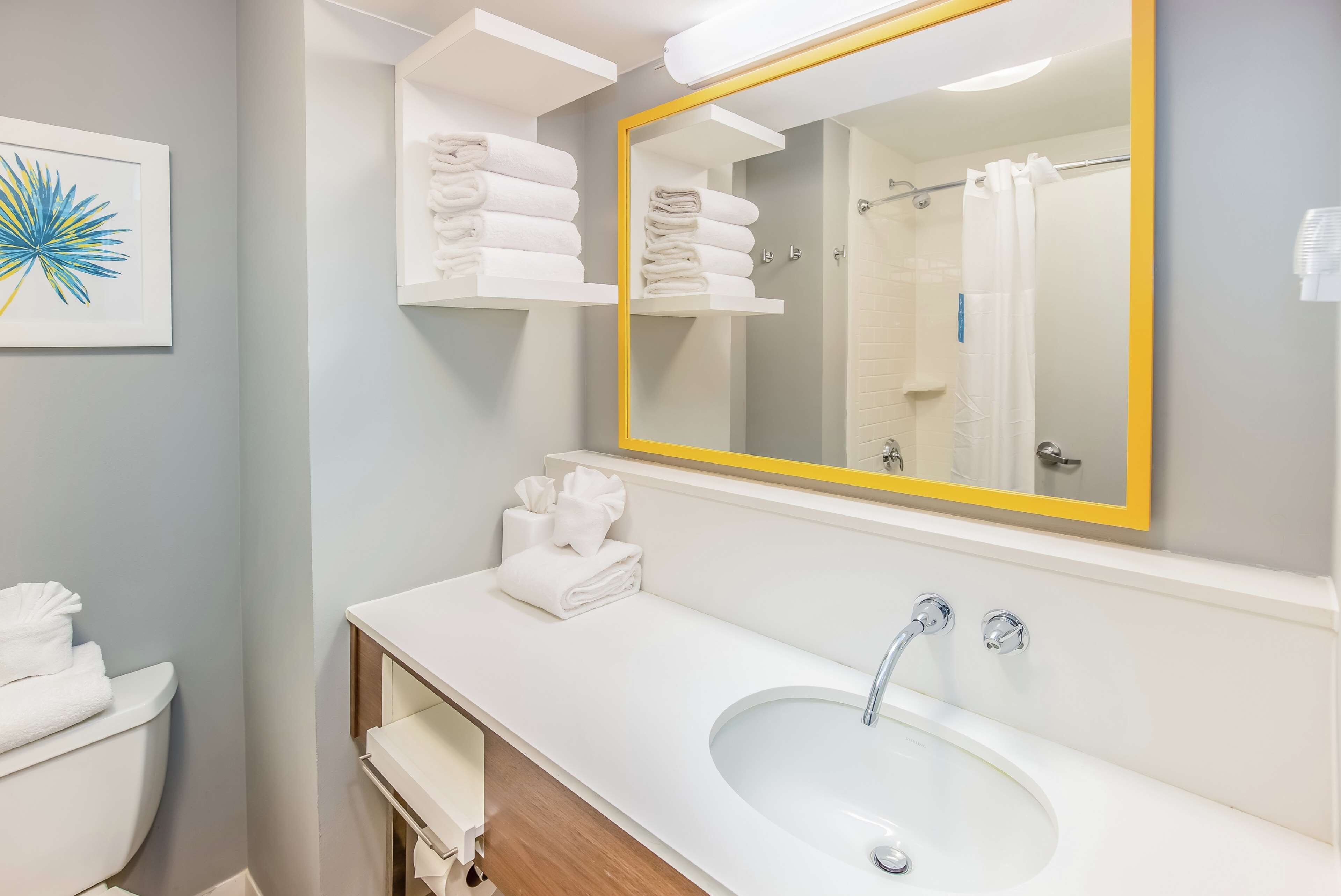 Un baño moderno con un lavamanos blanco, un espejo grande con marco amarillo y repisas con toallas blancas.