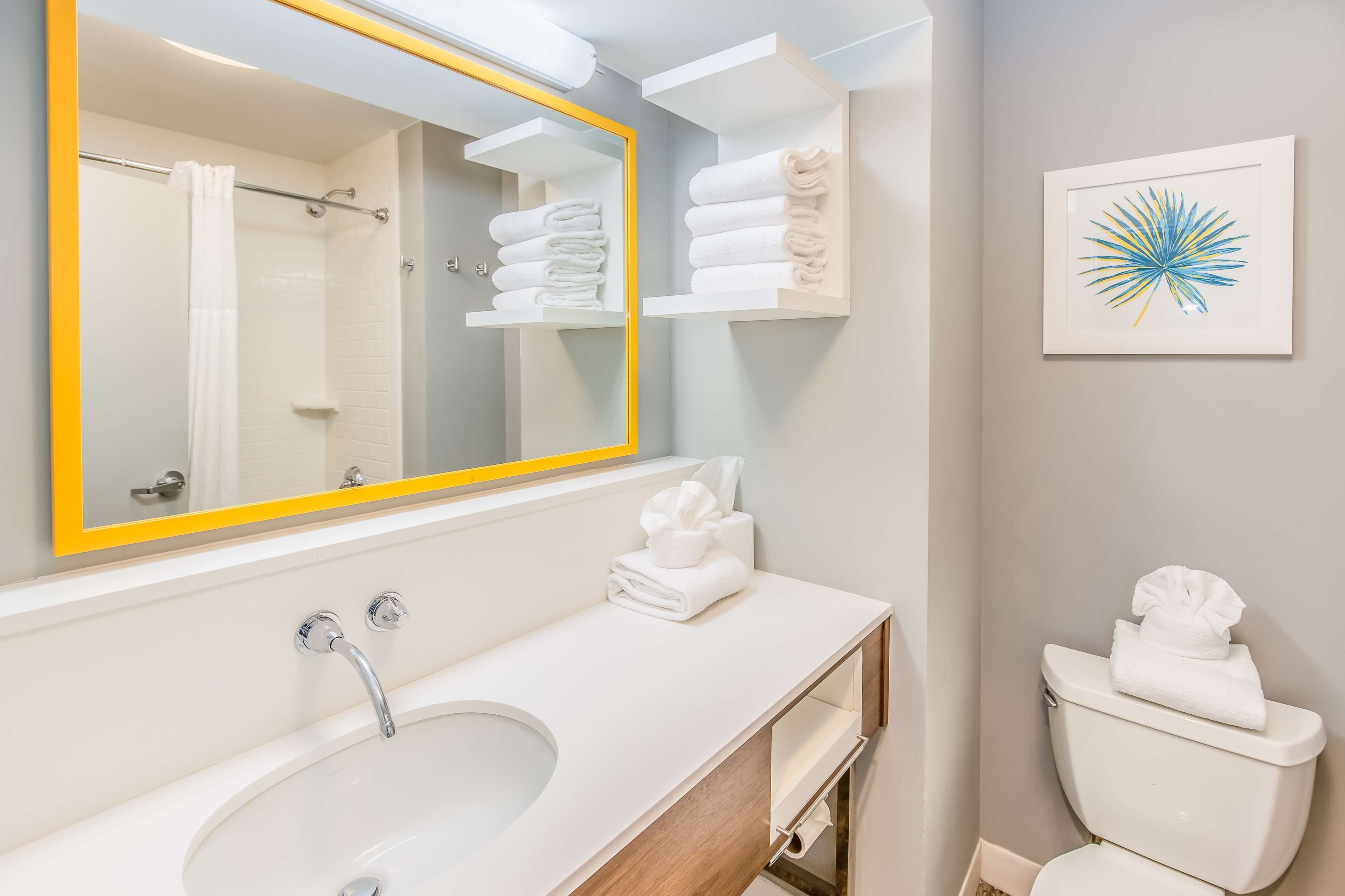 Une salle de bain moderne avec un lavabo blanc, un miroir avec un cadre jaune et des toilettes avec des serviettes.