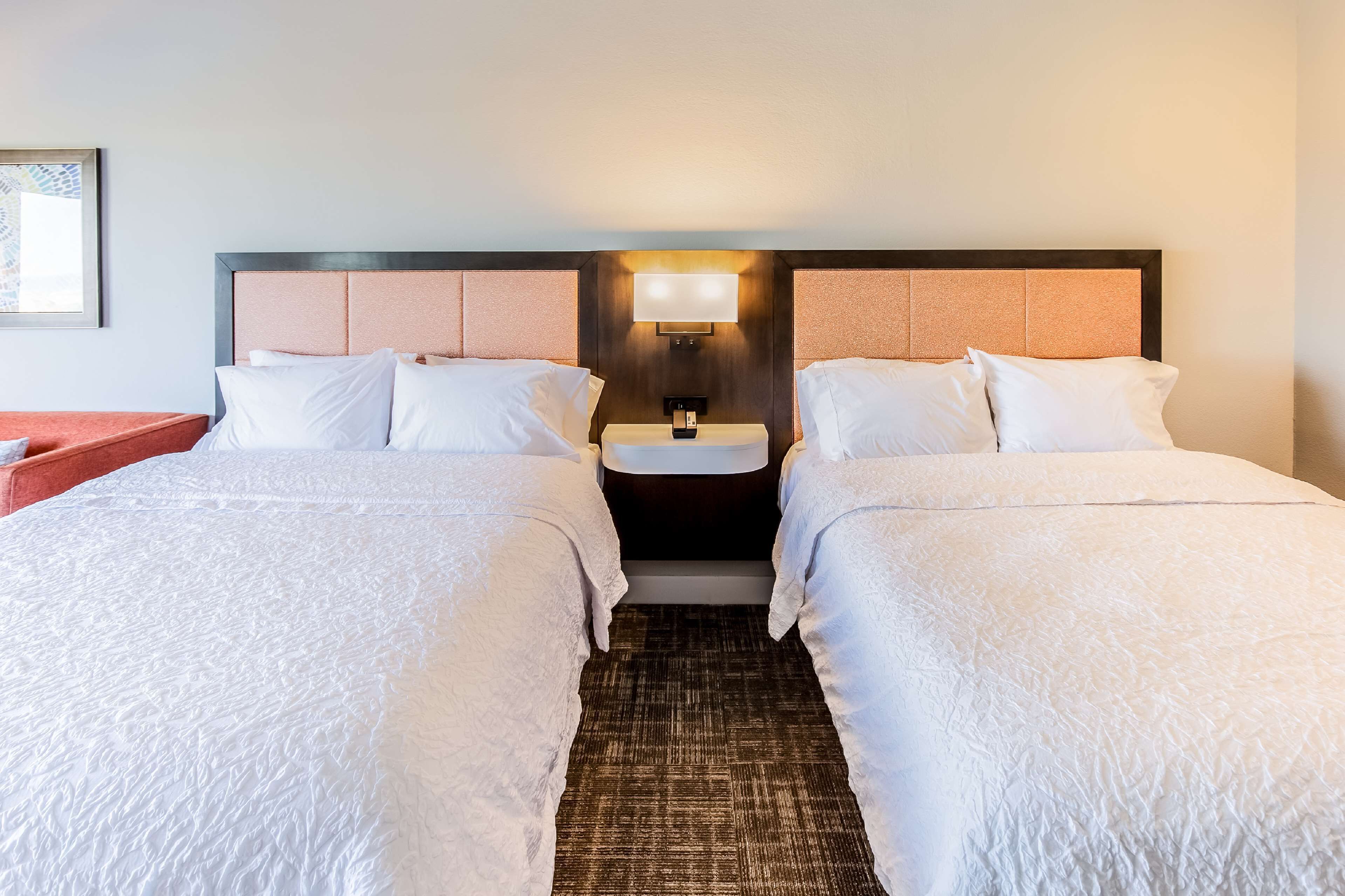 A hotel room with two beds, a nightstand, a wall-mounted lamp, and a small table.